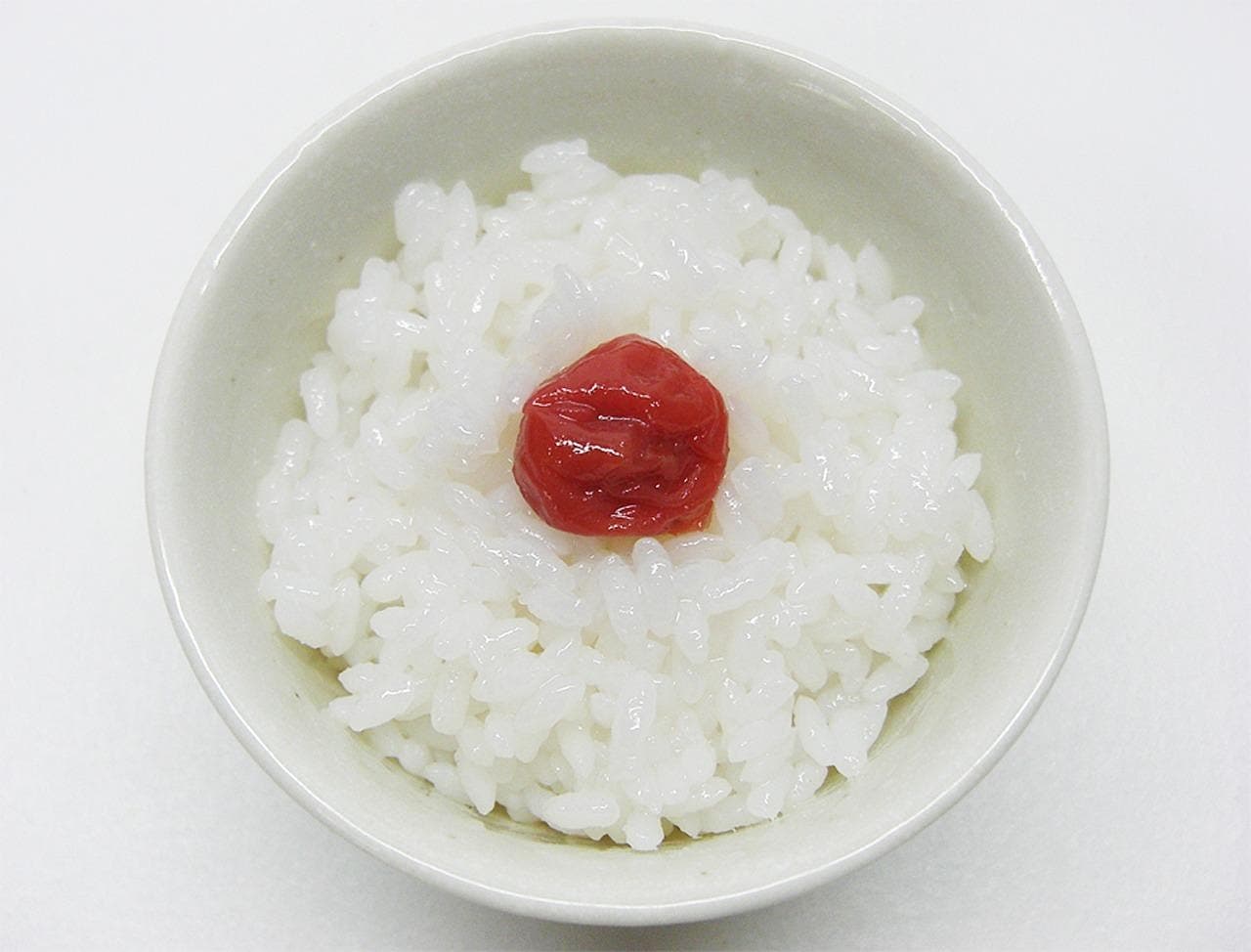 A white ceramic bowl of steamed white rice with a single red pickled umeboshi plum in the centre.