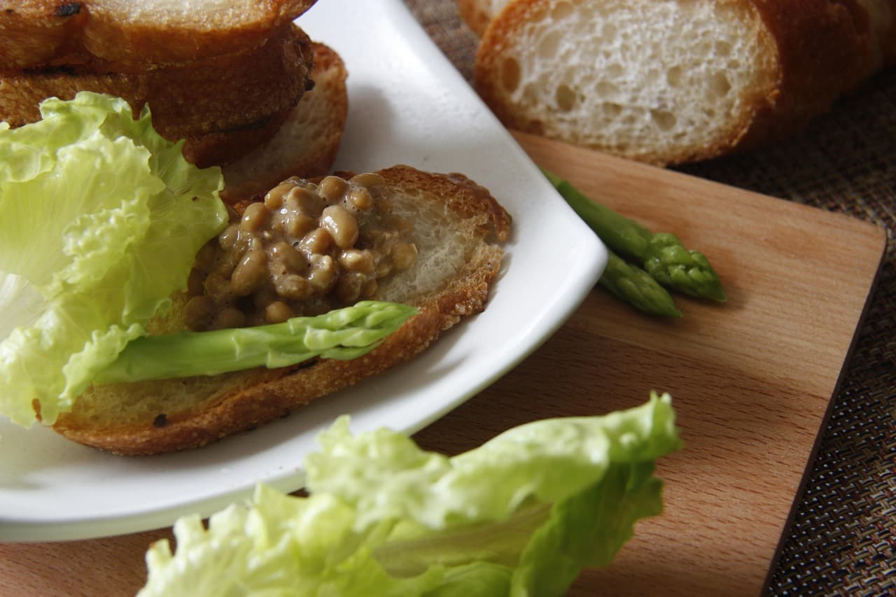 A slice of toasted baguette topped with sticky fermented natto beans and a spear of asparagus, served on a white plate with lettuce.