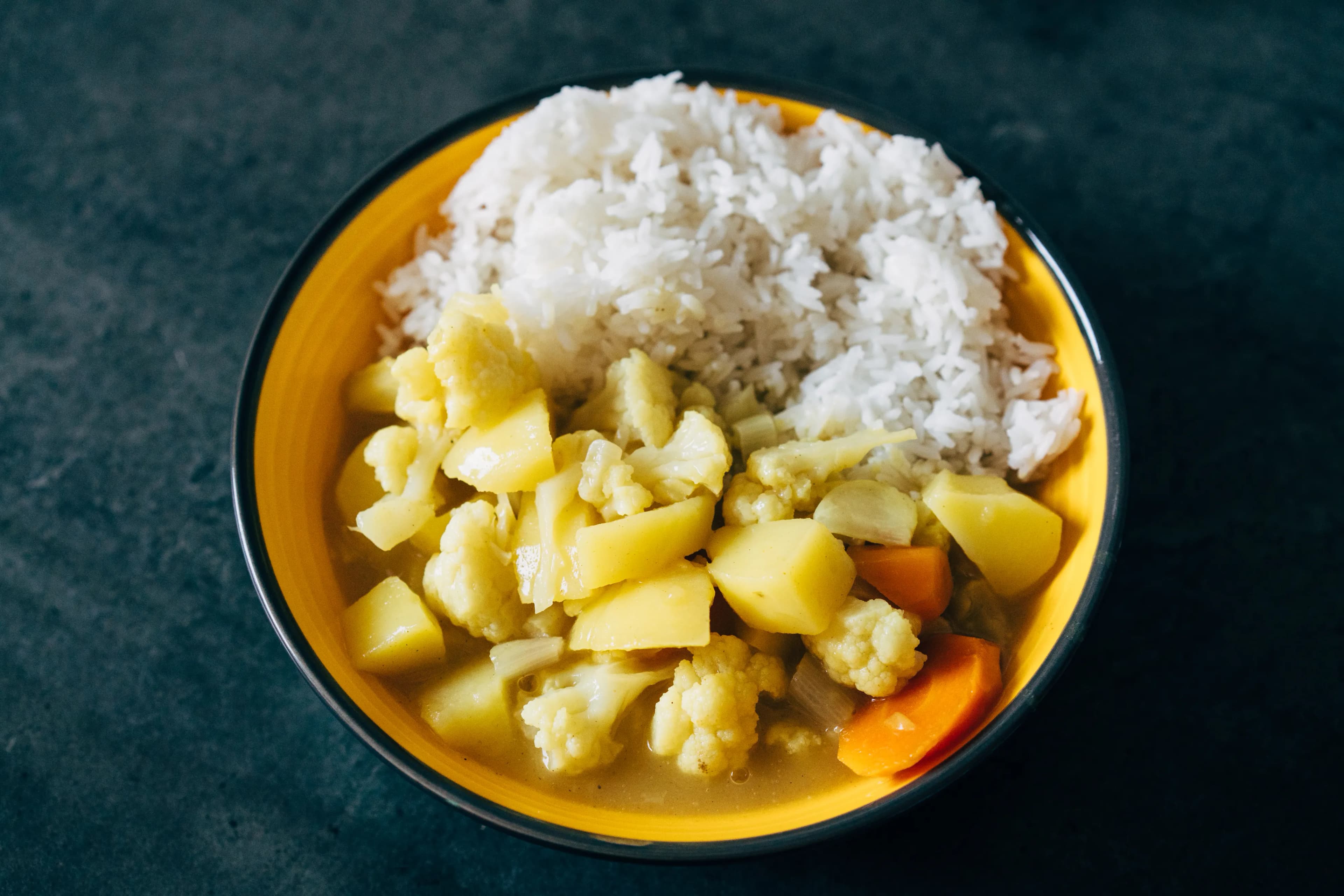 A yellow bowl of white rice with a vegetable curry of cauliflower florets, potato cubes and carrot slices in a yellow sauce, on a dark grey surface.