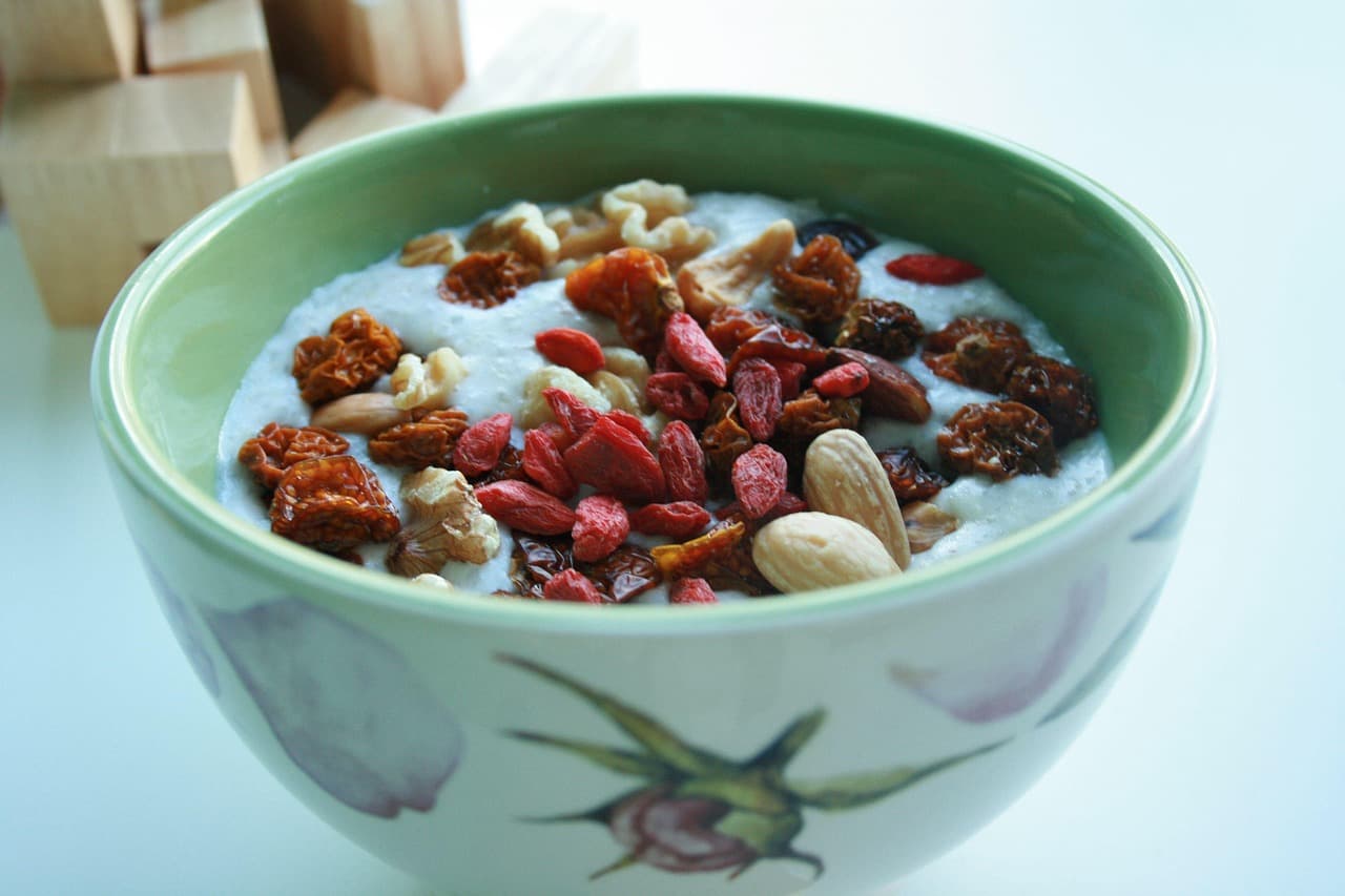 A bowl of pale oat porridge topped with red goji berries, whole almonds and walnut pieces, in a green and white floral bowl.