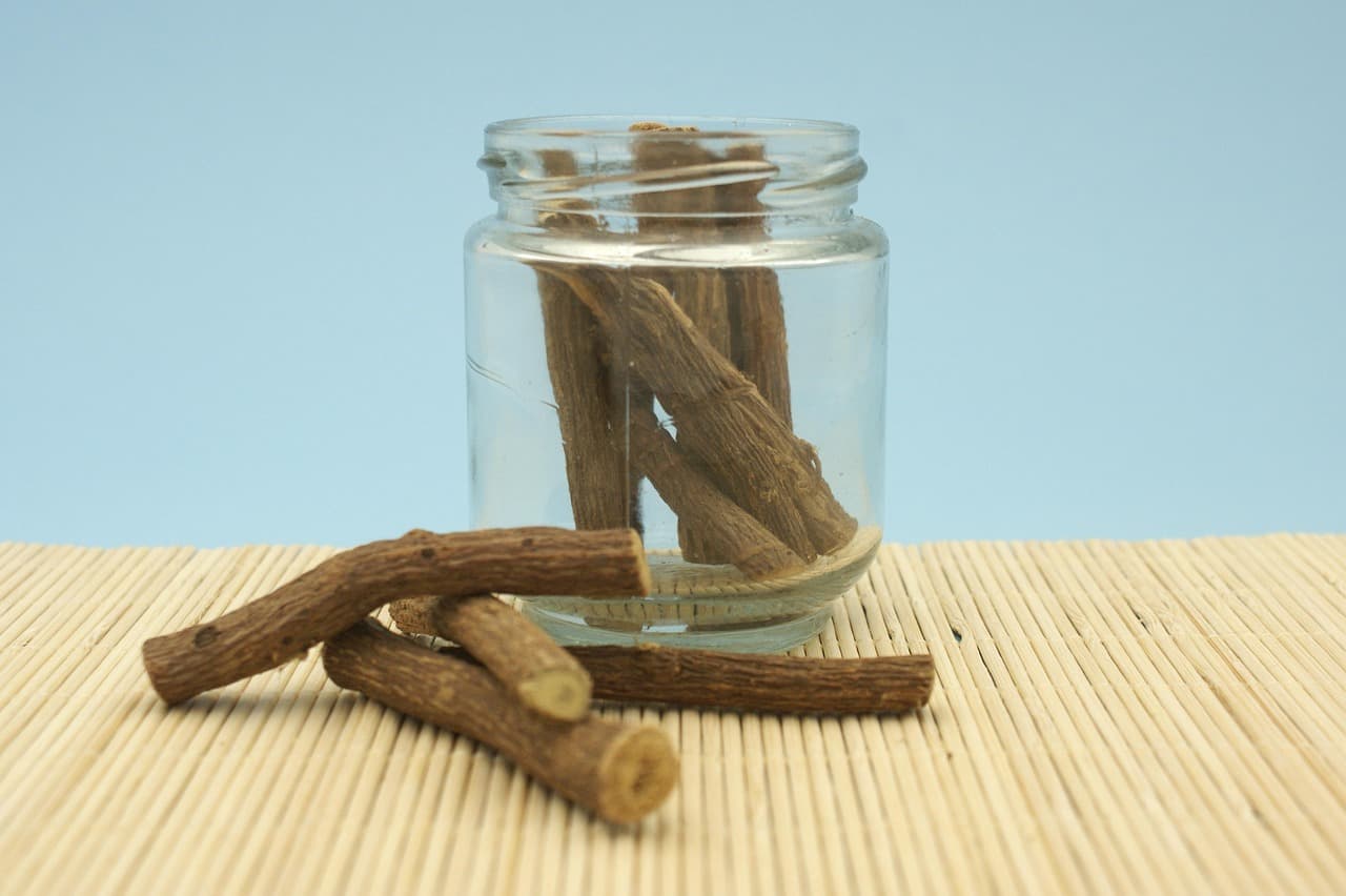 Dried sticks of licorice root standing in a small glass jar, with three more lying on a bamboo mat in front of it.